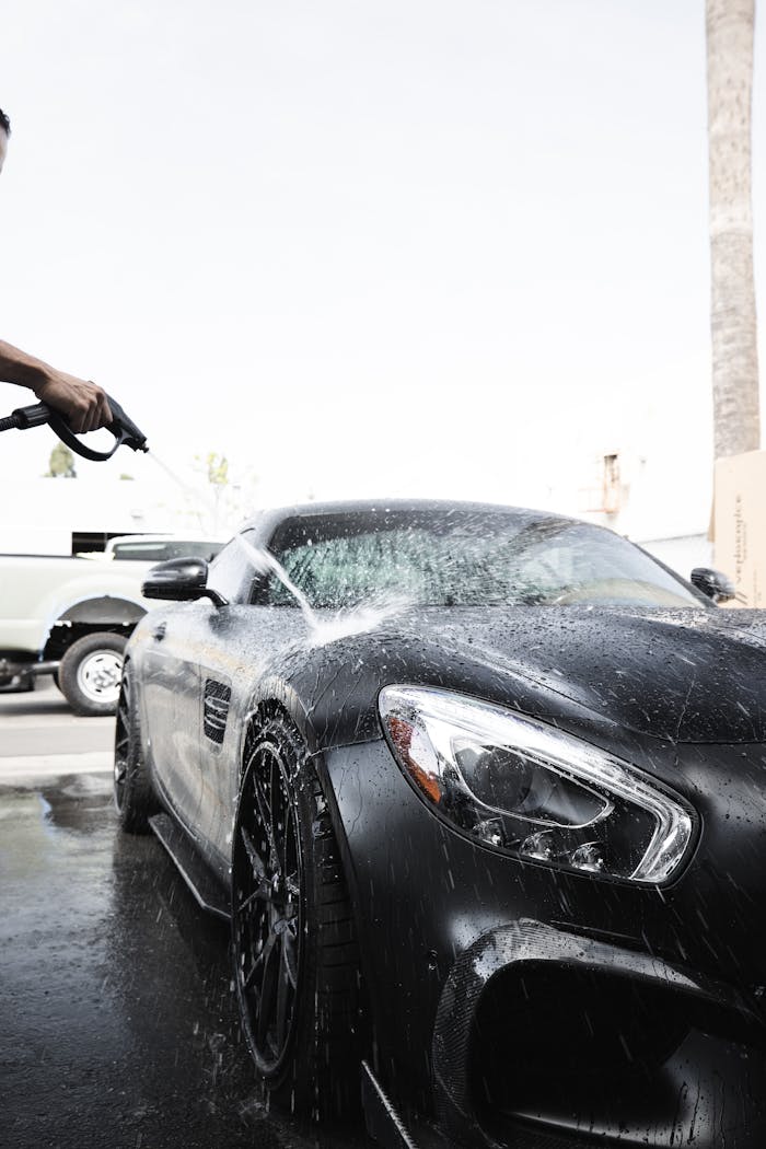 A luxury black car being washed with a pressure hose on a sunny day.