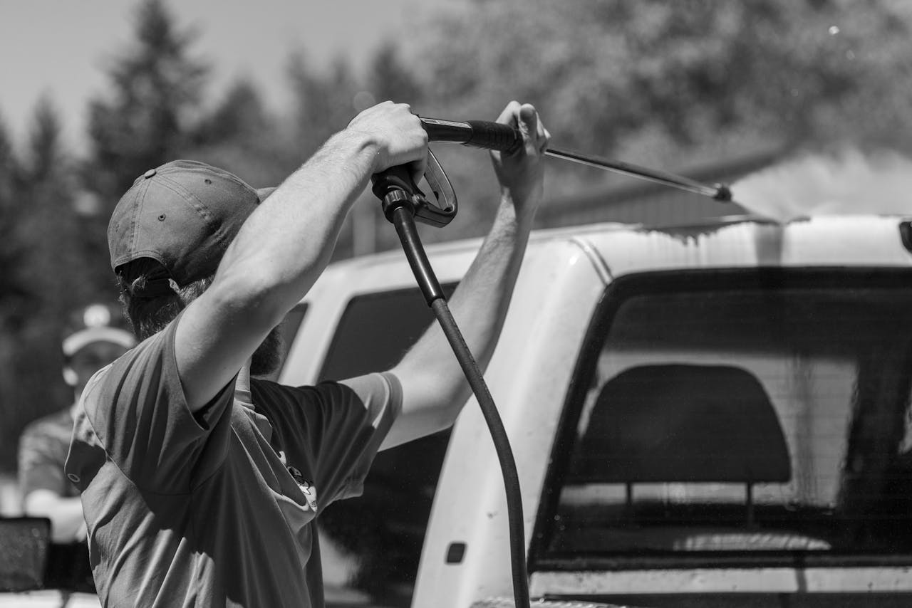 Home Black and white image of a man using a power spray on a truck outdoors.
